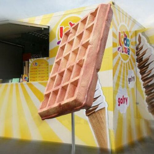 Large waffle stick statue in front of a yellow and white striped food stall decorated with ice cream cone graphics and the word