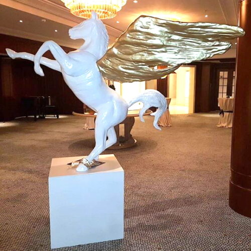 White Pegasus statue with golden wings displayed on a pedestal in a carpeted room with chandeliers.