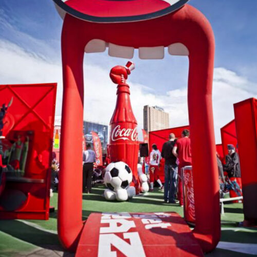 A large, inflated Coca-Cola-themed archway with giant teeth framing the entrance to a promotional event, featuring a large Coca-Cola bottle and a soccer ball sculpture. There are people milling around and red Coca-Cola banners in the background.