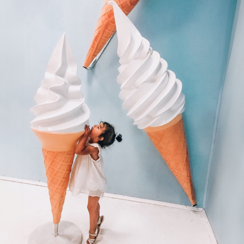 A child in a white dress standing and hugging a giant ice cream cone against a blue wall, with two more large ice cream cones attached to the wall in the background.