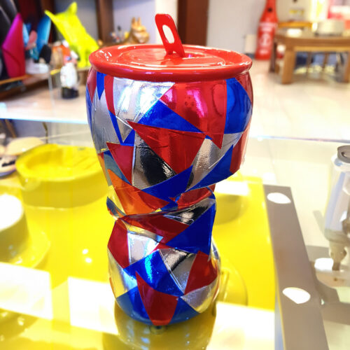 Colorful geometric-patterned ceramic vase with a red lid, placed on a reflective yellow surface.