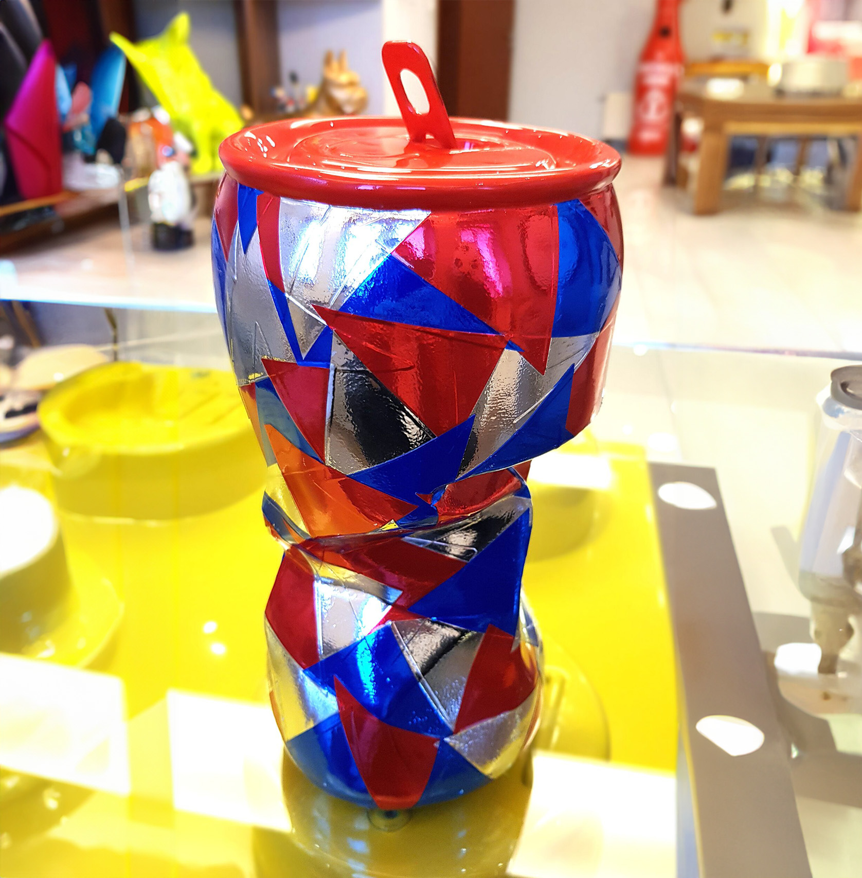 Pop art wrapping can, a functional and decorative figure. 2 Colorful geometric-patterned ceramic vase with a red lid, placed on a reflective yellow surface.