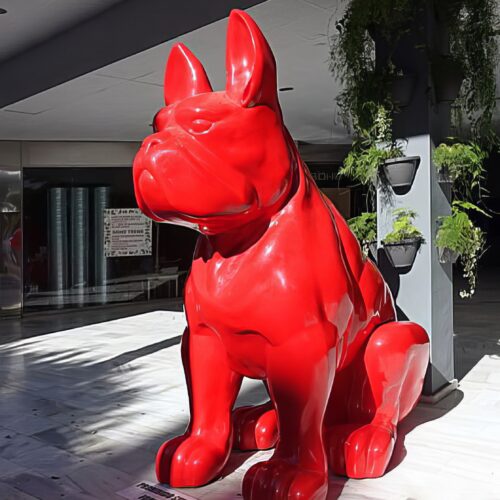 Large red sculpture of a sitting bulldog in an indoor area with potted plants on the side.