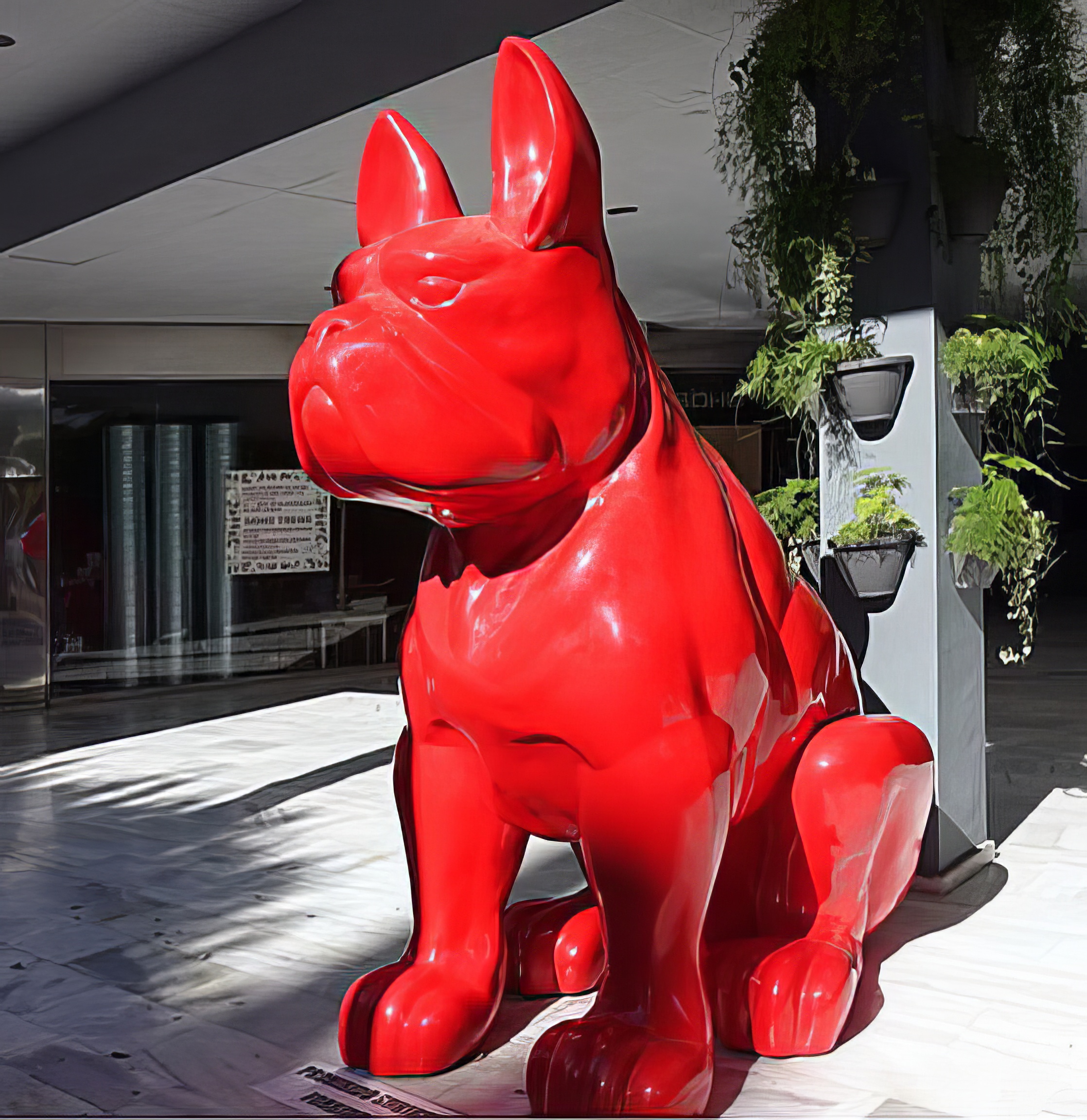 Giant sitting bulldog statue, single-colored. 2 Large red sculpture of a sitting bulldog in an indoor area with potted plants on the side.