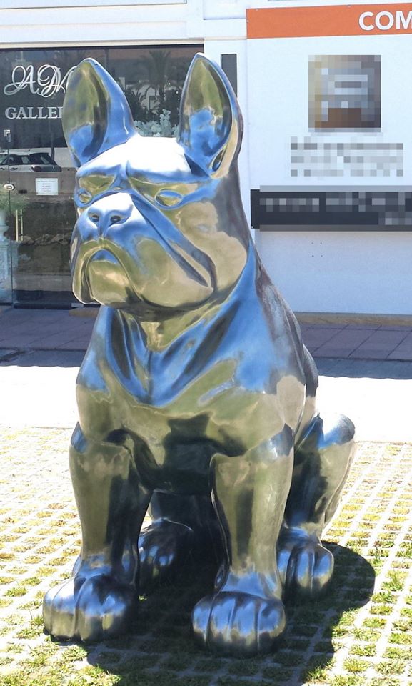 Giant sitting bulldog statue, single-colored. 4 Shiny metallic statue of a sitting dog with cropped ears, displayed outside a gallery.