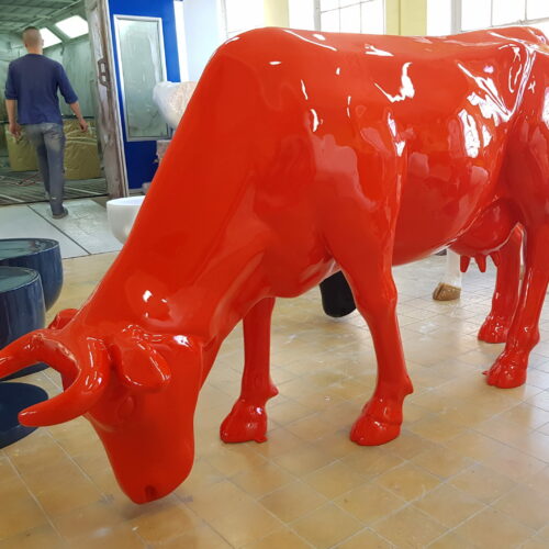 Red sculpture of a cow with its head lowered, positioned indoors with a person walking away in the background.