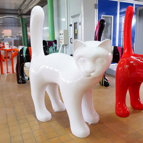 Large white cat sculpture with shiny surface, standing indoors among other colorful abstract animal sculptures.