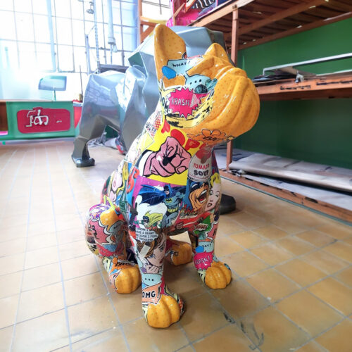 Colorful bulldog sculpture decorated with various comic book images and pop art designs, sitting on a tiled floor in an indoor space with shelves and a large window in the background.