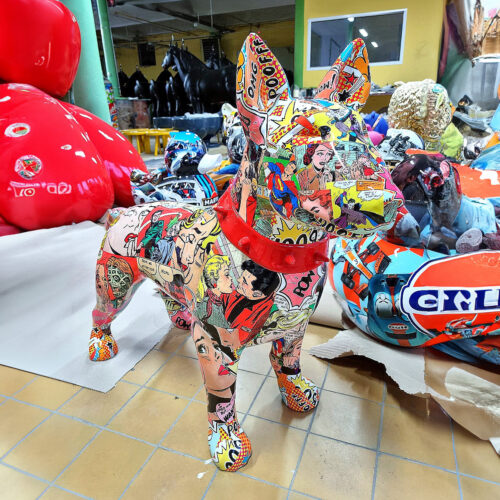 A dog statue covered in colorful comic book art, featuring a red collar and set in a room filled with various large, vibrant sculptures, including a large red figure and black horse statues in the background.