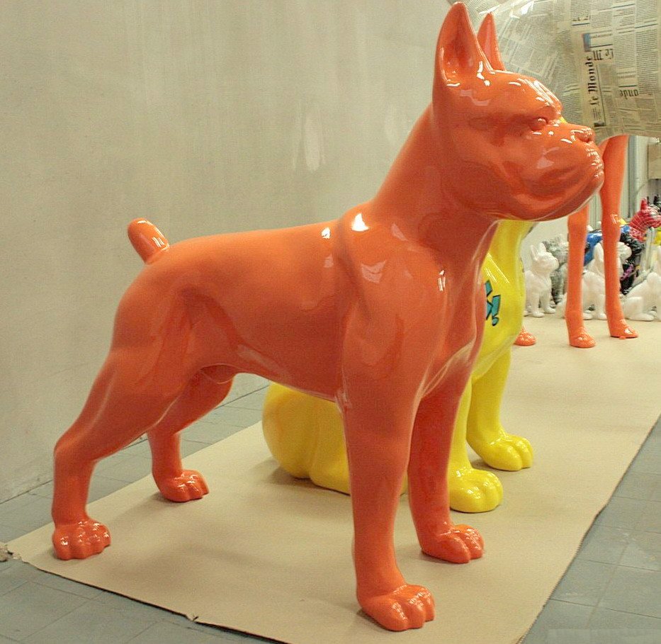 Boxer Dog Figure Standing - Single Color 2 Large orange dog sculpture with a shiny surface standing on a beige platform, with partial views of yellow and white dog sculptures in the background.