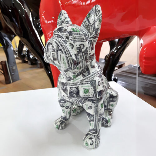 A sculpture of a French Bulldog covered in dollar bill designs, placed on a white surface with other art pieces in the background.