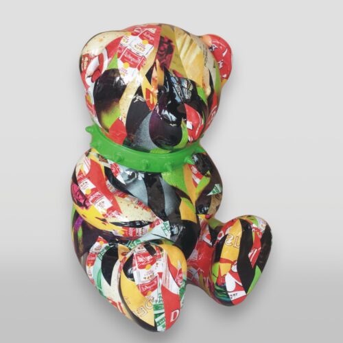 A teddy bear sculpture made from colorful, collaged paper materials sits with a bright green collar against a white background.