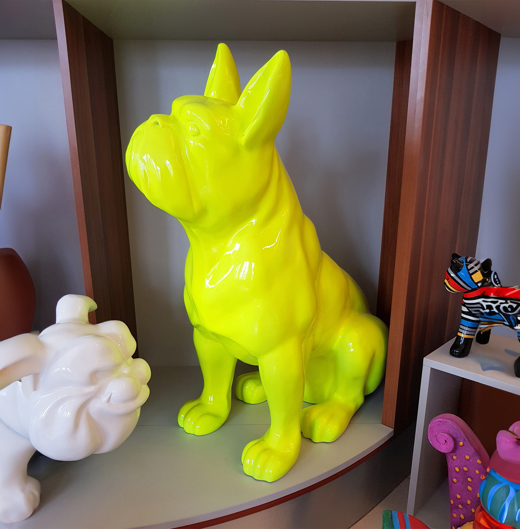 Middle size seated bulldog 1 Bright yellow statue of a sitting French Bulldog displayed on a shelf among other colorful sculptures.