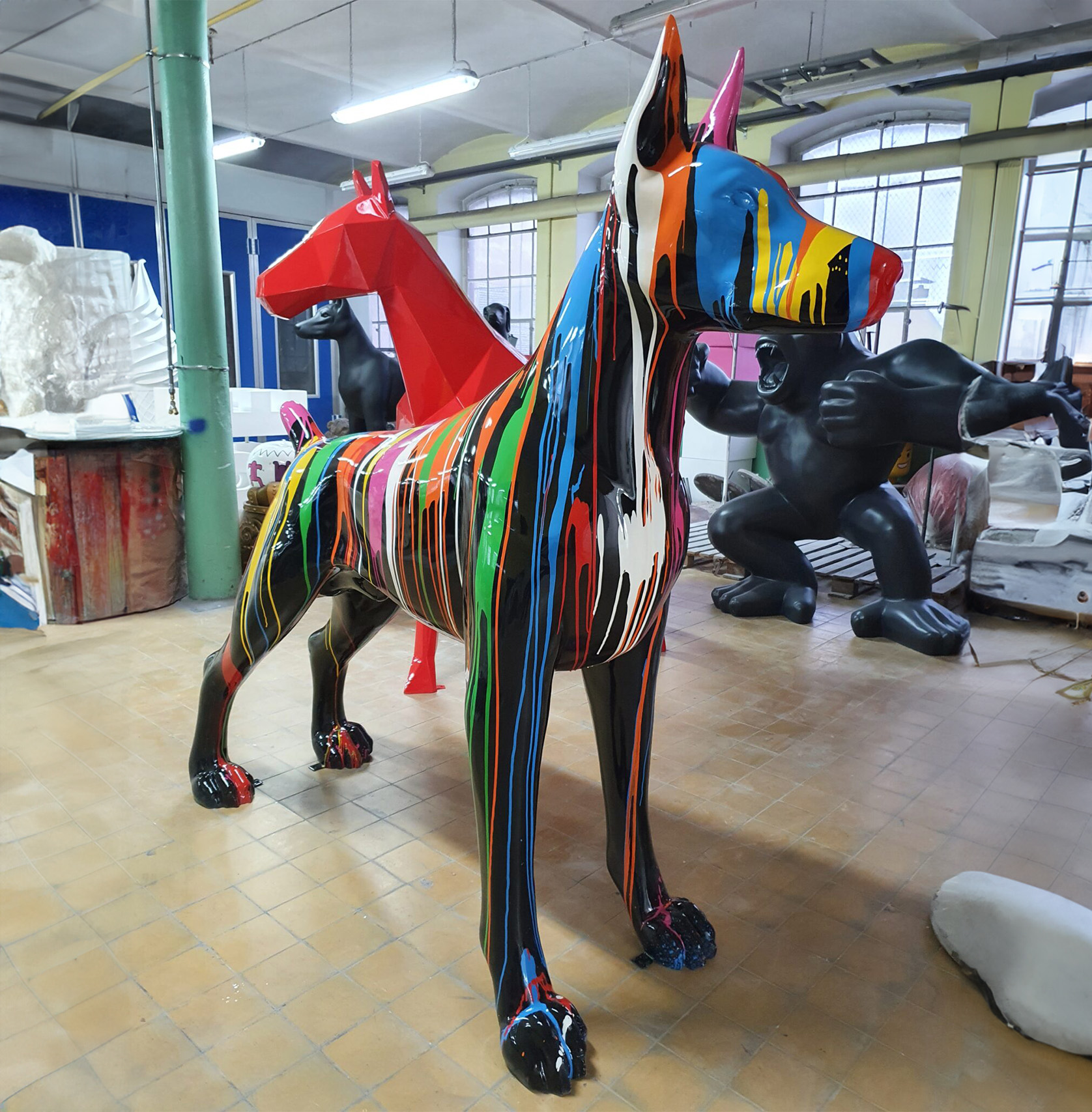 Large Doberman Colorful Fiberphglass Statue 1 Colorful life-sized sculpture of a dog with multicolored paint drips, inside an art studio with other large sculptures in the background.