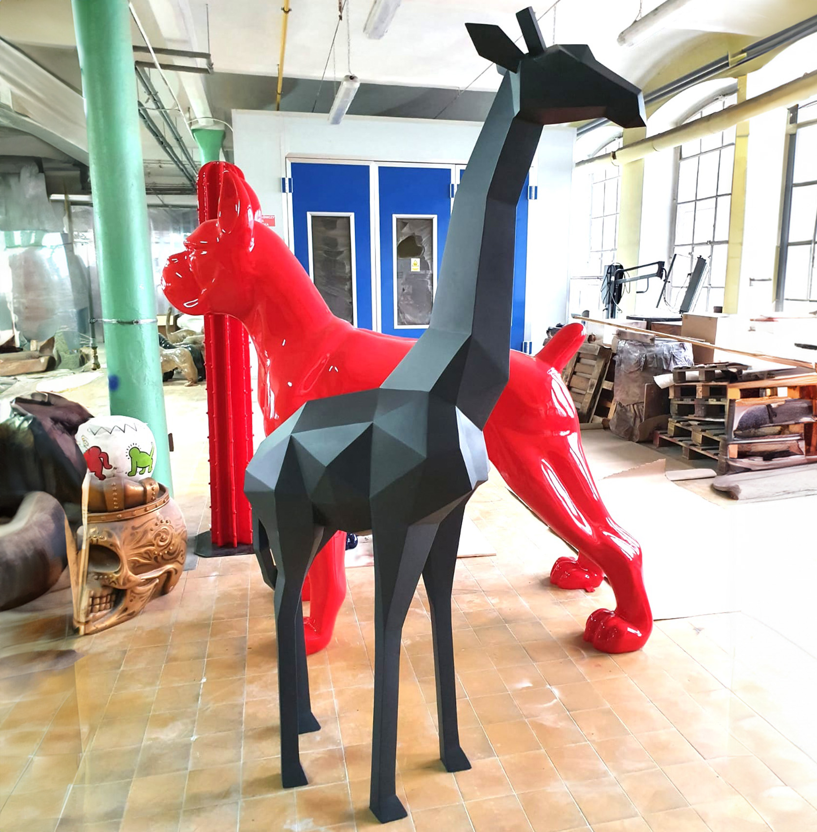 Large geometric fiberglass giraffe statue 2 Geometric black giraffe sculpture in front of a glossy red dog sculpture, in a workshop setting with tools and materials in the background.