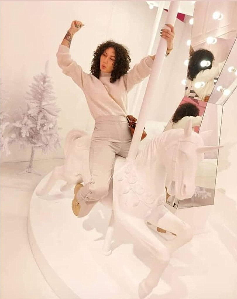 Carousel of unique unicorns 3 Osoba siedząca na karuzeli z białym jednorożcem w jasnym pomieszczeniu, obok znajdują się lustra i ozdobione białe choinki.