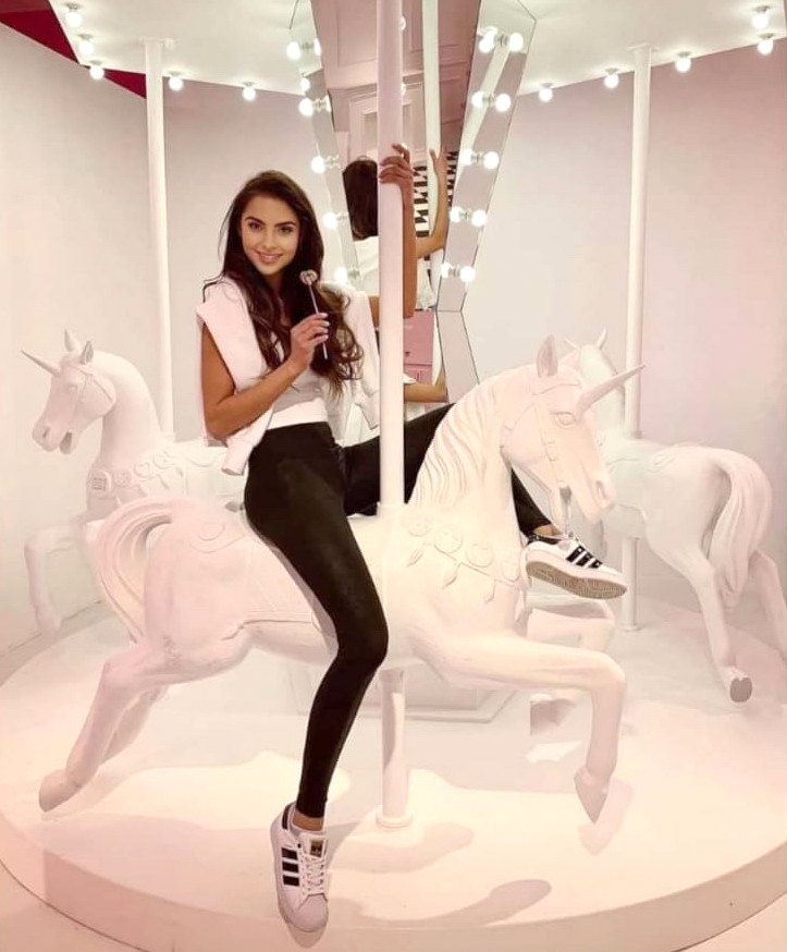 Carousel of unique unicorns 1 Kobieta siedząca na białym jednorożcu na karuzeli, trzymająca lizaka, otoczona jasnymi światłami.