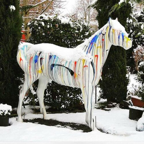 A white horse statue with multicolored drip paint design, covered in snow, set in a snowy garden with greenery in the background.