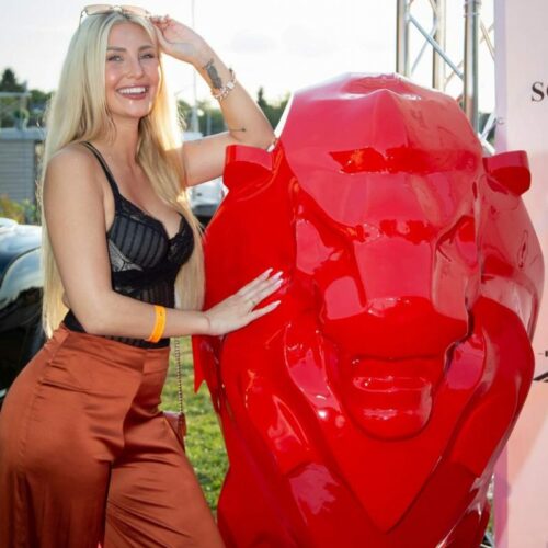 A woman with long blonde hair and wearing a black top and brown pants stands next to a large red lion statue, smiling and holding sunglasses atop her head.