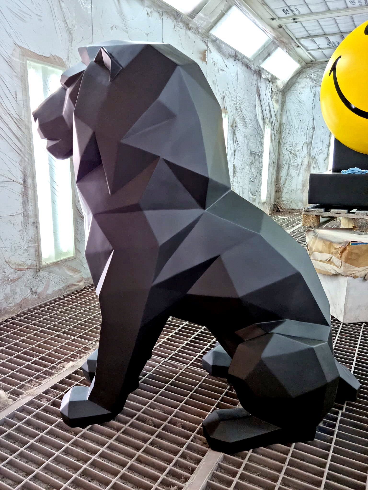 Monochromatic Geometric Lion Figure 4 Black polygonal lion sculpture in a textured, industrial workshop setting with a large yellow smiley face sphere partially visible in the background.