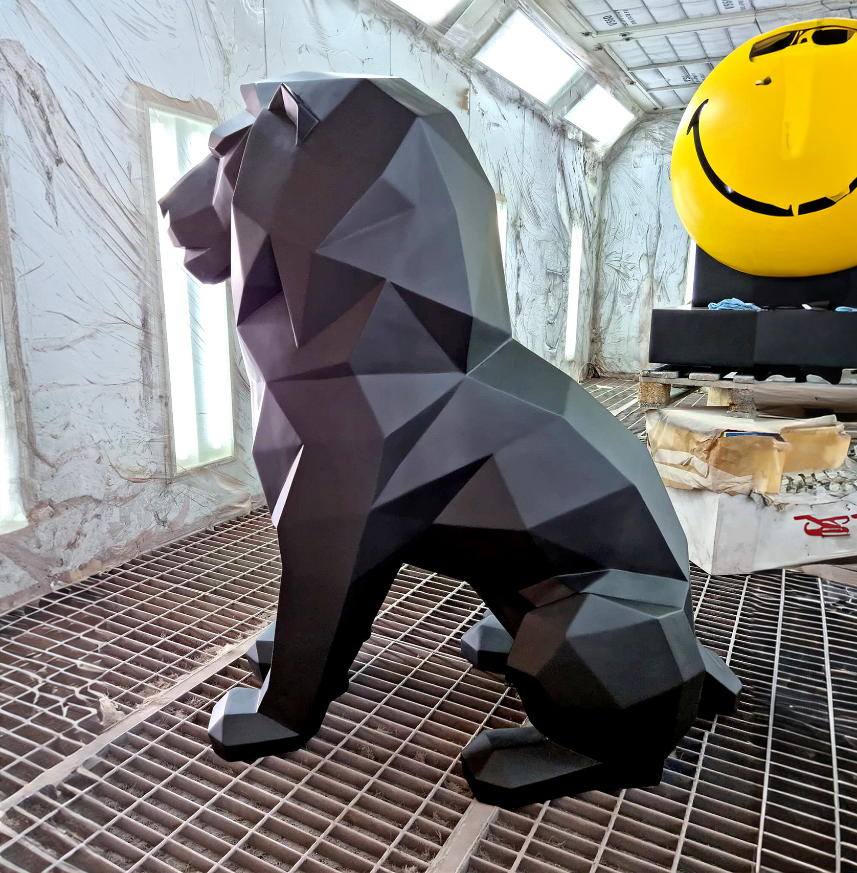 Geometric lion sculpture single color 2 Geometric black lion sculpture inside a workshop with a yellow smiley face sculpture in the background.