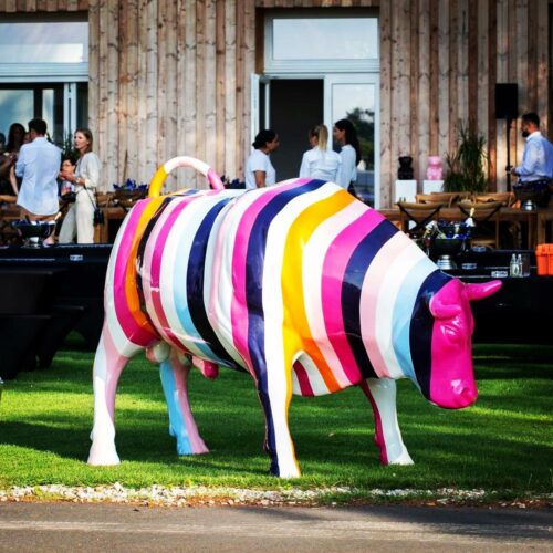 A large colorful cow sculpture with vertical stripes in various colors stands on a lawn with people socializing in the background.
