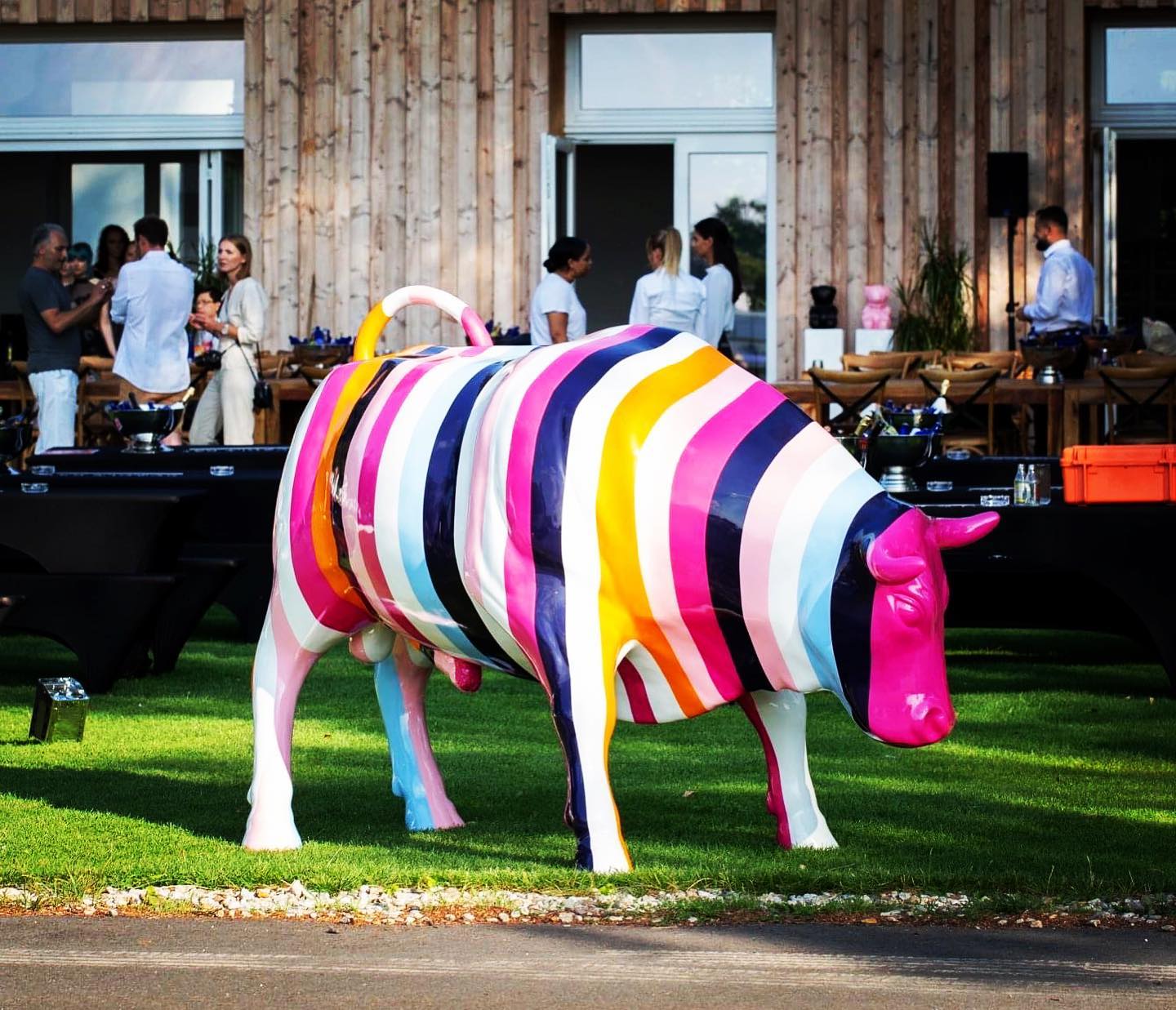 Striped fiberglass large bull statue 1 A large colorful cow sculpture with vertical stripes in various colors stands on a lawn with people socializing in the background.