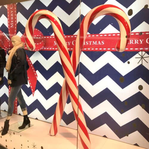 Christmas window display with two large candy canes and a mannequin wearing a scarf against a blue and white chevron background.