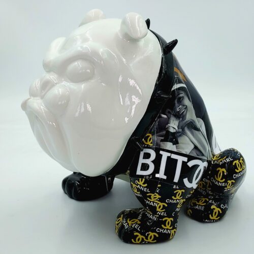 Bulldog-shaped sculpture with a glossy white face, black spike collar, and legs wrapped in black material featuring yellow Chanel logos. The body has a collage of black and white photos and the text 
