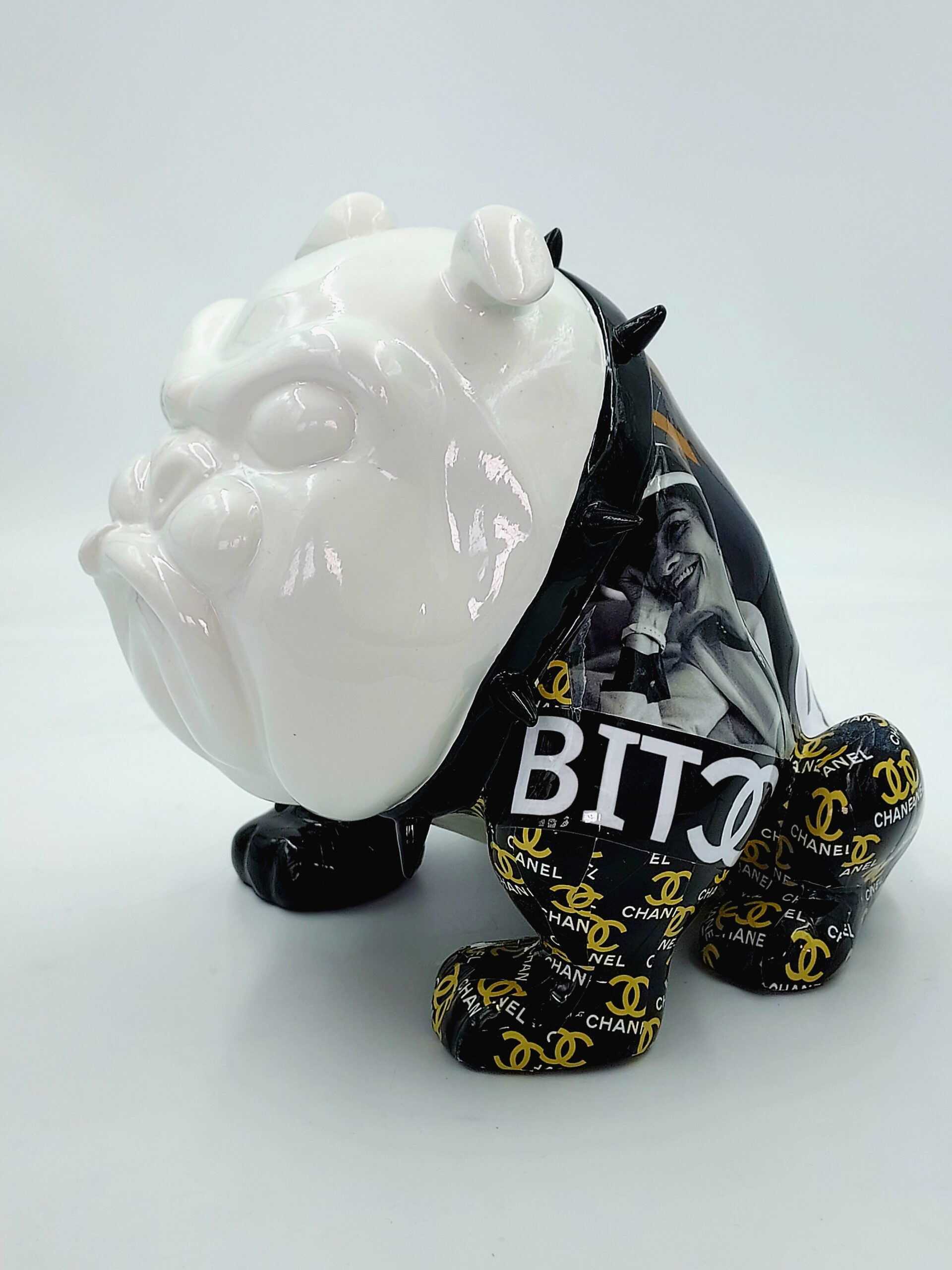 Bulldog Avella 55cm - Vogue 1 Bulldog-shaped sculpture with a glossy white face, black spike collar, and legs wrapped in black material featuring yellow Chanel logos. The body has a collage of black and white photos and the text