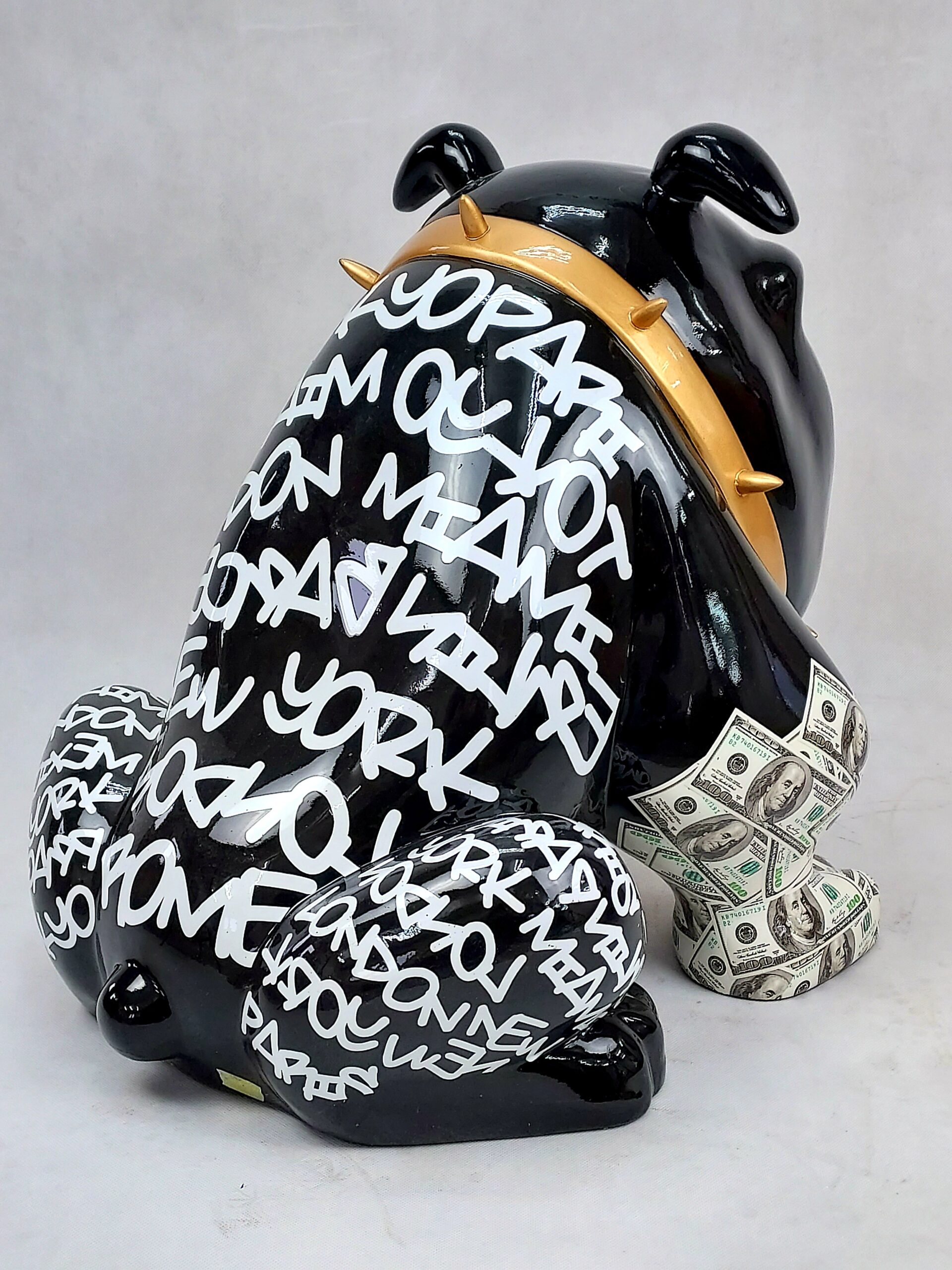 Bulldog Avella 55cm dog - Louis Paris 3 Black bull-shaped sculpture with gold spikes around the neck, covered in white graffiti-style text of city names, holding a bundle of U.S. dollar bills.