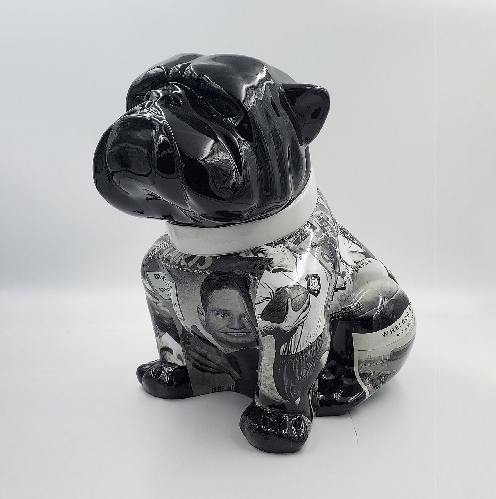 Bulldog Angelo 40cm dog - rugby 1 Sculpture of a sitting bulldog with a shiny black head and neck, and a body decorated with monochrome images and text clippings.