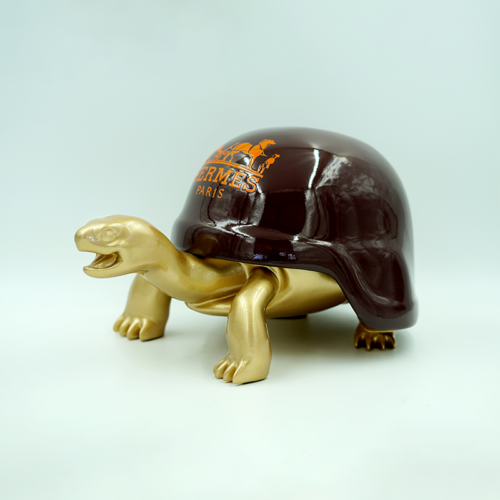 Gold turtle sculpture with dark brown shell, featuring the Hermès Paris logo in orange on the shell.