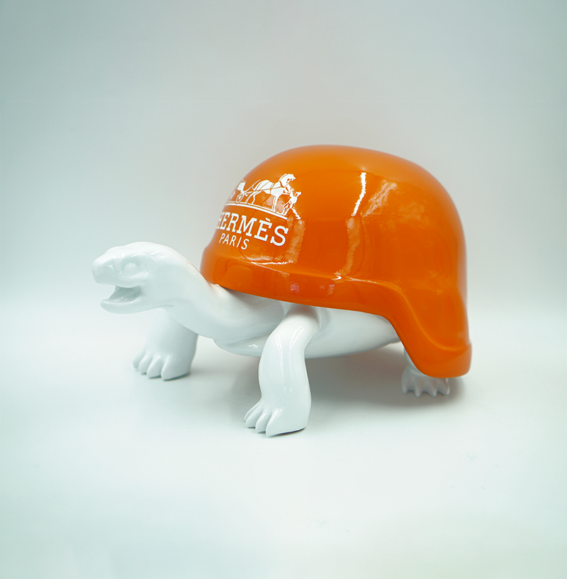 Turtle in a Helmet - H 1 A white ceramic turtle figurine with an orange shell featuring the Hermès Paris logo.