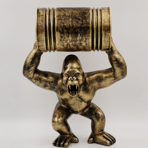 A bronze-colored statue of a roaring gorilla standing on two feet, holding a barrel above its head with both hands.
