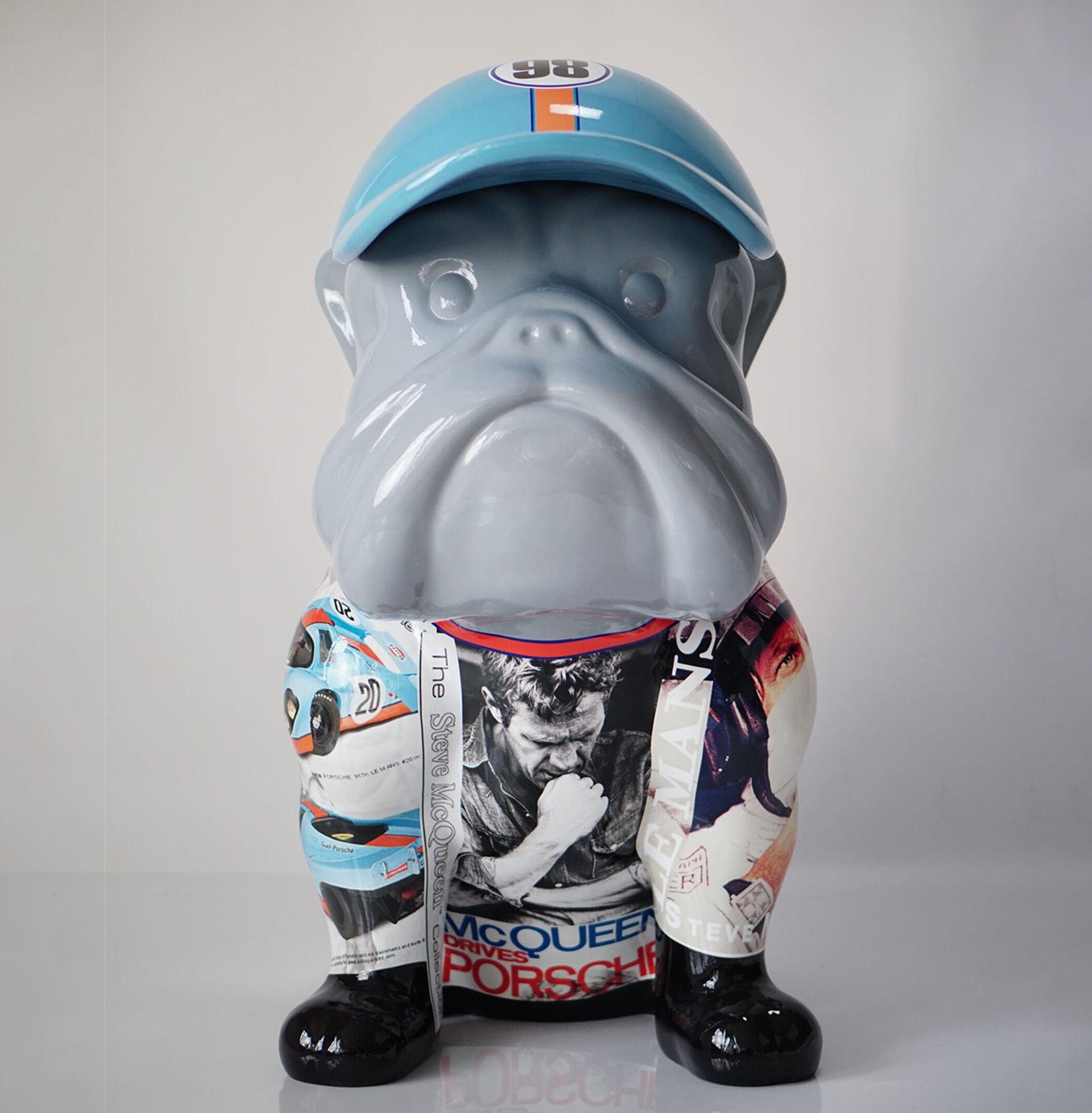 Bulldog dog with hat - McQueen 2 Szara figurka psa w niebieskim kapeluszu, ozdobiona kolorowymi zdjęciami samochodów wyścigowych i portretami ludzi.