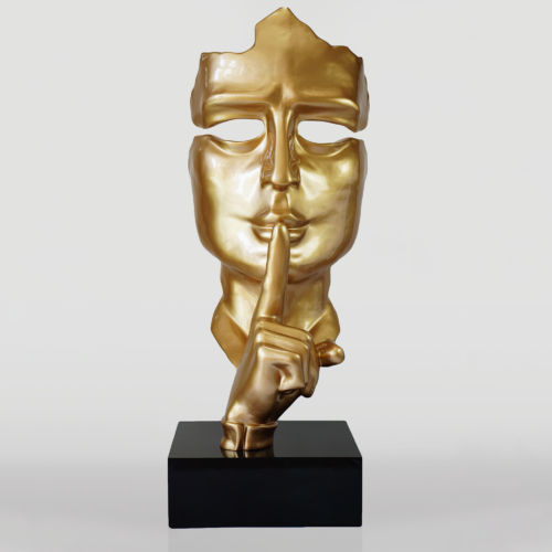 Gold sculpture of a face with a finger pressed against its lips, symbolizing silence, placed on a black base.