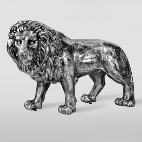 A metallic sculpture of a lion with a detailed mane, standing on all four legs against a light grey background.