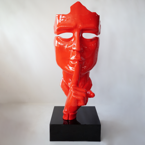 Red sculpture of a human face with a missing top half, pressing a finger to its lips, mounted on a black base.