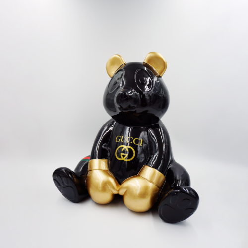 A black glossy teddy bear sculpture with gold accents on its ears, hands, and feet, featuring the