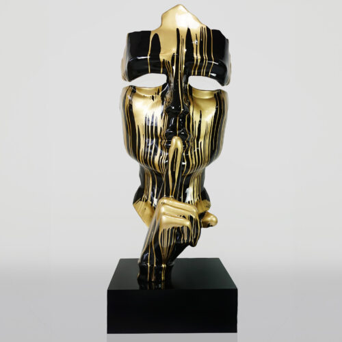 Rzeźba z maską w kolorach złotym i czarnym, z palcem na ustach, umieszczona na czarnym postumencie.