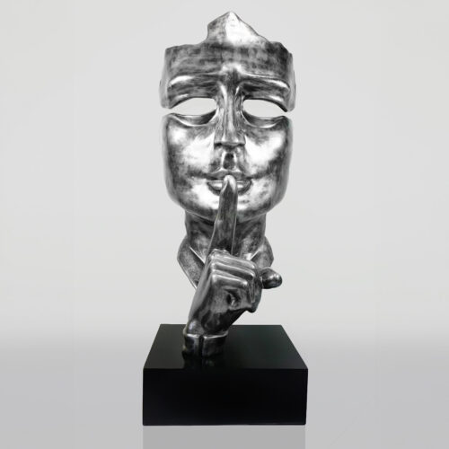 Silver sculpture of a masked face with a finger held up to its lips in a shushing gesture, mounted on a black base.