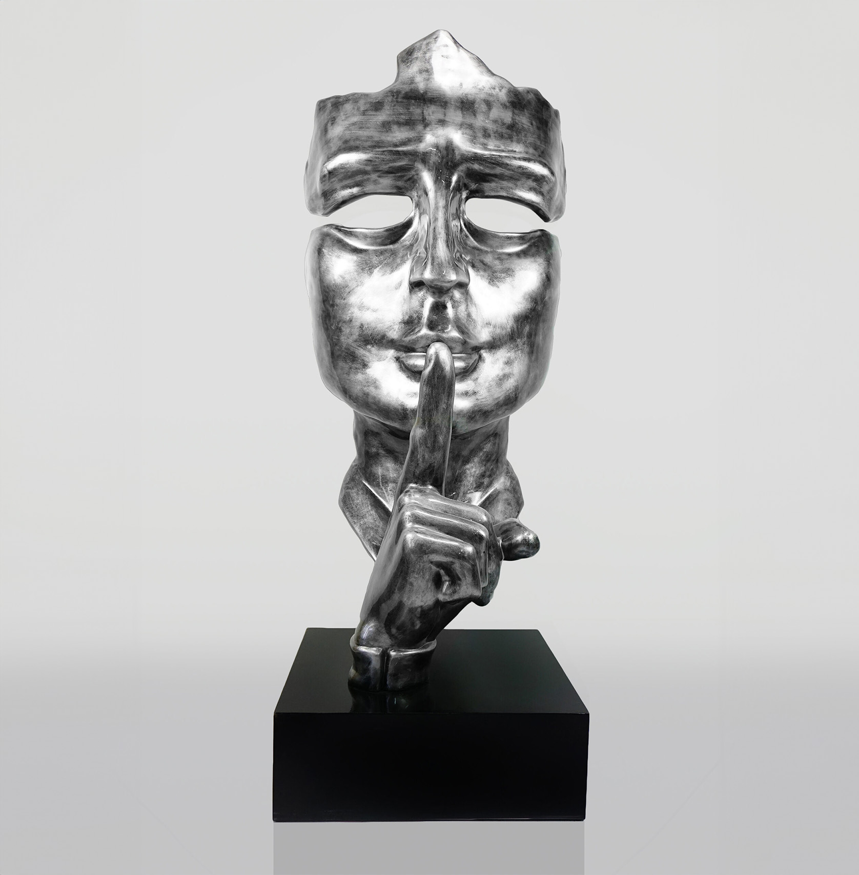 Large mask on stand - Silver Patina 1 Silver sculpture of a masked face with a finger held up to its lips in a shushing gesture, mounted on a black base.