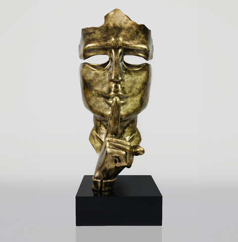 Golden sculpture of a partially abstract face making a shushing gesture, mounted on a black rectangular base.