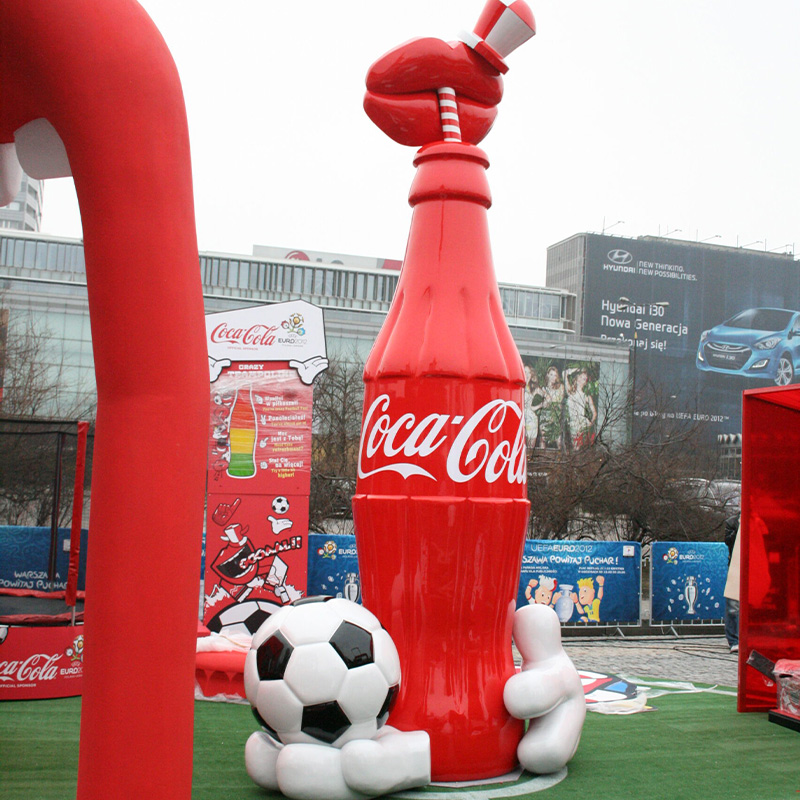 oryg_064490685_1129949550544944_1465385417682452480_o Large inflatable Coca-Cola bottle with a red, white, and gray cap, and a white gloved hand holding a soccer ball at the base, displayed in front of a building and advertising banners.
