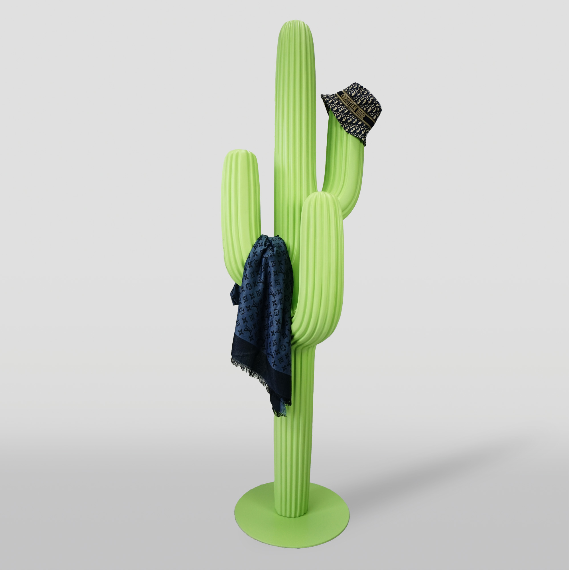 Pastel green hanger - cactus 2 A light green cactus-shaped coat rack with a dark blue scarf draped over one arm and a black patterned hat hanging on another arm, standing on a circular base against a plain background.
