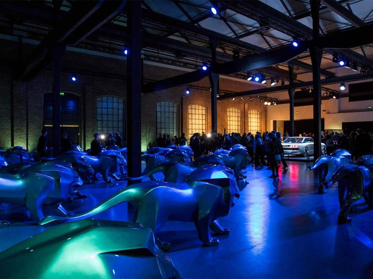 Peugeot lion fiberglass advertising sculpture 2 Pomieszczenie wypełnione niebieskimi rzeźbami lwów, z ludźmi i samochodem w tle, oświetlone niebieskim i fioletowym światłem.