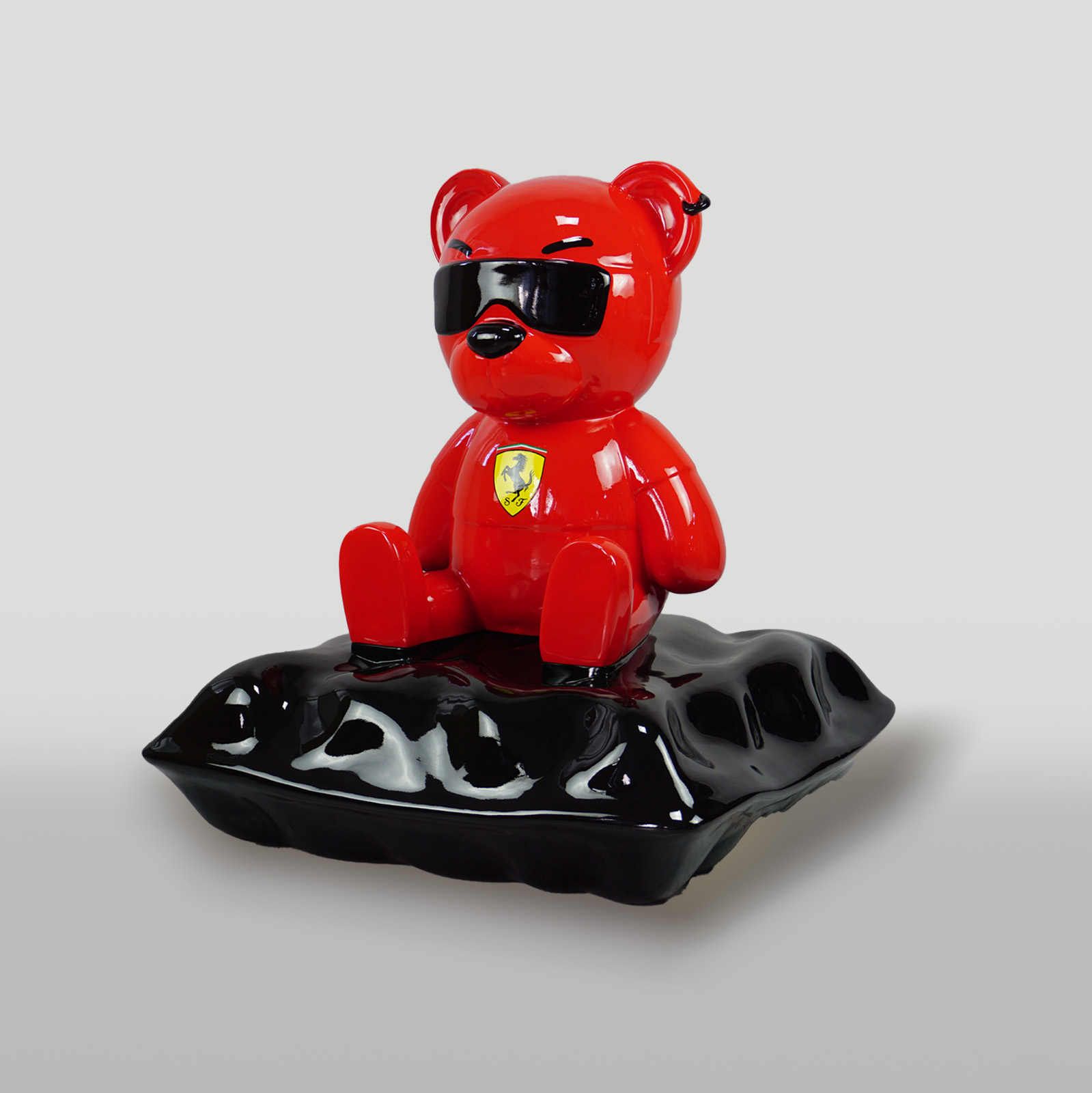 A red bear figurine wearing black sunglasses and a Ferrari logo, sitting on a glossy black surface.