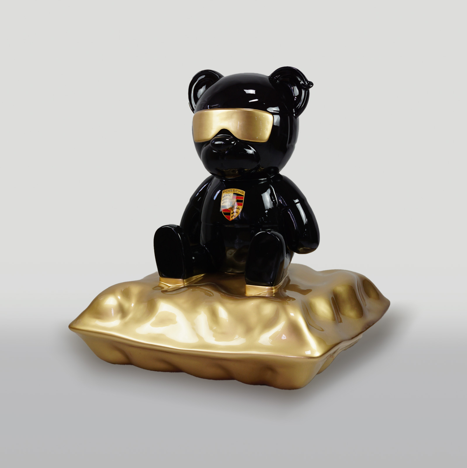 Porsche Teddy Bear sitting on a pillow 1 A black bear statue with gold sunglasses, seated on a plush gold pillow, featuring a Porsche logo on its chest.