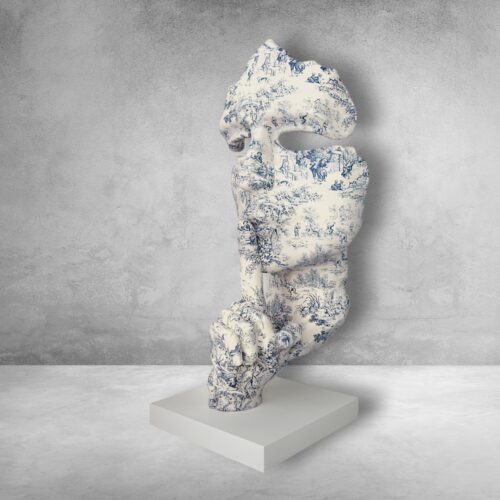 A sculpture resembling a stylized human figure decorated with intricate blue and white patterns, positioned on a plain white pedestal against a textured grey background.